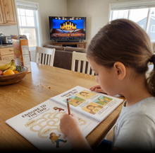 Load image into Gallery viewer, Child coloring a book at a dining table with a TV in the background
